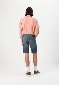 Kortærmet, lyserød skjorte med en tekstureret overflade, parret med blå denimshorts. Modellen har brune sko og hvide sokker på.