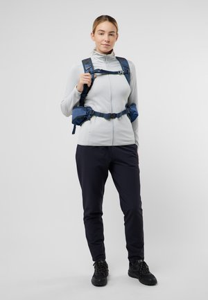 Giacca zip con cappuccio grigio chiaro, pantaloni scuri e scarpe nere. Zaino blu con spallacci regolabili e una funzione di supporto in vita integrata.