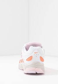 Baskets Nike blanches et roses vues de dos, avec des détails orange et une semelle beige texturée sur une surface blanche.