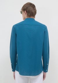 Camicia a maniche lunghe color turchese con una texture morbida, polsini abbottonati e un design con yoke sul retro. Presenta un colletto classico e una vestibilità rilassata.