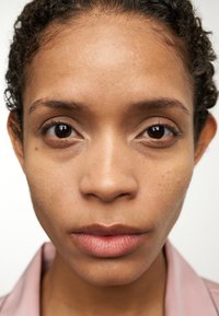 Nahaufnahme eines Gesichts mit mittlerem Hautton, definierten Zügen, dunklem lockigem Haar und neutralem Make-up; trägt ein hellrosa Hemd mit Kragen.