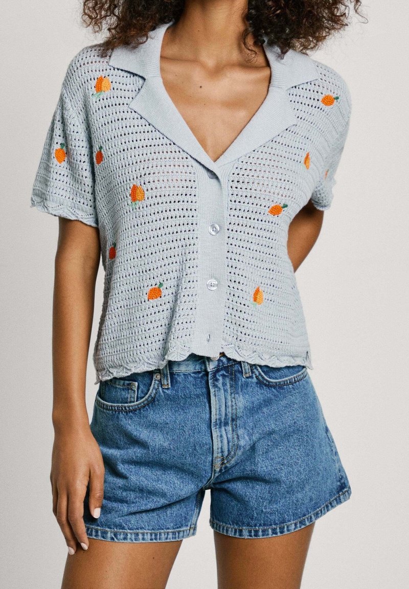 Cardigan all'uncinetto azzurro chiaro con ricamo di frutti arancioni, chiusura a bottoni e colletto a scollo; abbinato a pantaloni di jeans.