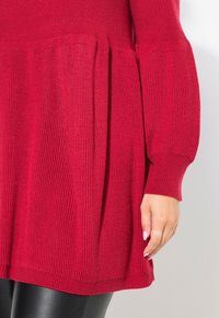 Rote Strick-Tunika mit gerippter Textur, die ein ausgestelltes Design und lange Ärmel hat und oberhalb der Hüften endet. Darunter sind schwarze, lederähnliche Hosen sichtbar.