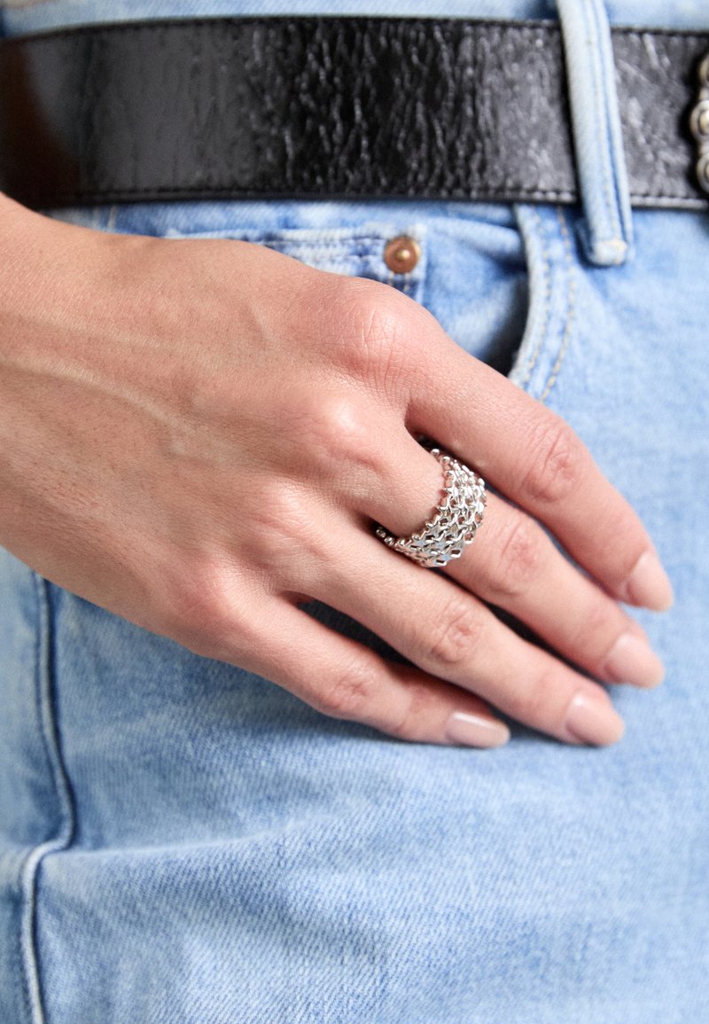 Silver multi-band ring with interlocking links, displayed on a hand against a light denim background. Features smooth texture and shiny finish.