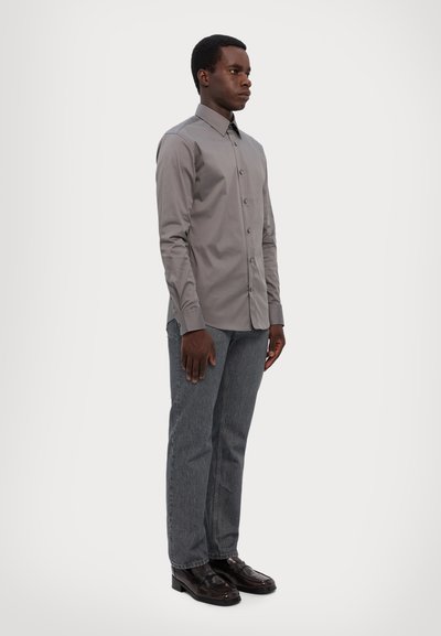 Chemise gris clair à manches longues avec boutons et col, associée à un jean gris foncé et des chaussures noires. Le tissu semble lisse et ajusté.