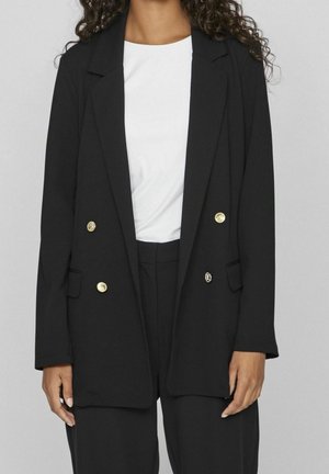 Kobieta ubrana w czarną marynarkę dwurzędową z złotymi guzikami, na białej koszuli i czarnych spodniach na jednolitym tle.