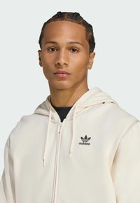 Sudadera con cremallera de color beige claro que cuenta con capucha con cordones, bolsillos frontales y un logo de trébol negro de Adidas en el pecho. Textura suave, ajuste casual.