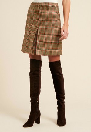 Woman wearing a knee-length brown and red plaid skirt paired with black suede over-the-knee heeled boots, standing against a plain background.