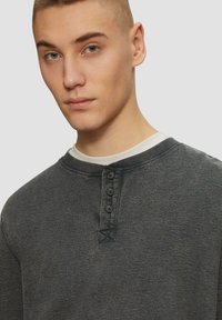 Tumehall pika varrukaga henley-s�ärk, millel on neli esiknappi, ümmargune kaelus ja tekstureeritud kangas. Valge aluspluus on nähtav kaeluse juures.