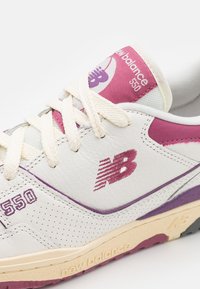 Fehér bőr sportcipő rózsaszín és lila akcentussal. Perforációkkal és vastag fűzőkkel rendelkezik. "New Balance 550" logóval a nyelven és az oldalon.
