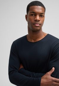 Dunkelblaues Langarmshirt aus weichem Stoff. Verfügt über einen Rundhalsausschnitt und eine figurbetonte Silhouette. Die Arme des Modells sind verschränkt.