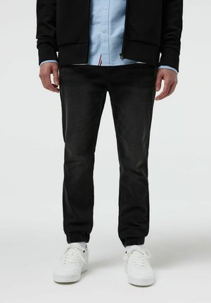 Osoba ubrana w czarne spodnie dresowe, białe trampki, jasnoniebieską koszulę na guziki oraz czarną kurtkę na zamek, stojąca z rozluźnionymi rękami.