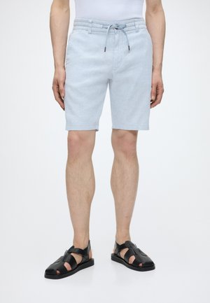 Férfi, világoskék zsinóros rövidnadrágban és fekete pántos bőrszandálban, fehér, egyszínű háttér előtt áll.