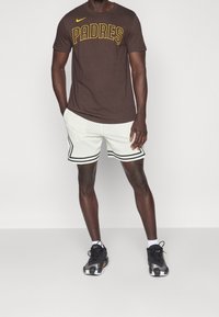 Brun Nike t-shirt med "PADRES" text i gult, parat med vita mesh shorts med svart kant och svarta träningsskor.