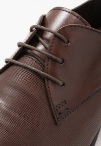 Hnědá kožená bota s texturovaným povrchem, s hladkou částí na svršku, kulatými tkaničkami a švy poblíž ok řetězů.