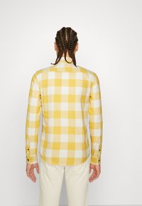 Camisa de cuadros amarillos y blancos, de botones, con mangas largas y puños, hecha de una tela ligera, que presenta un dobladillo curvado y un yugo trasero.