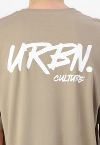 Tricou khaki dintr-un material moale, cu un text mare alb pe spate care citește "URBN. CULTURE.", având o croială lejeră și guler clasic.