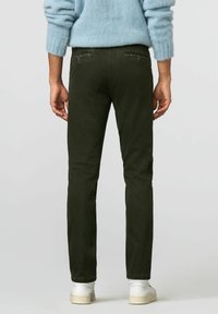 Donkergroene slim-fit jeans met achterzakken met decoratieve stiksels, gezien van achteren. De stof heeft een gladde textuur. Witte sneakers zichtbaar.