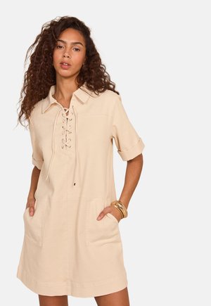 Vrouw met krullend haar die een beige jurk met korte mouwen, veterkraag en zakken draagt, staand met haar handen in de zakken.