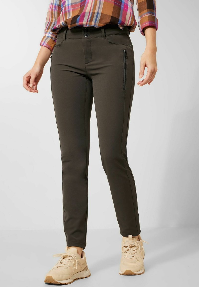 Femme portant un pantalon slim vert olive foncé avec une poche zippée sur la cuisse, des baskets beiges et une chemise à carreaux multicolores rentrée.