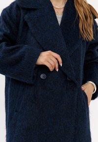 Donna che indossa un cappotto a doppio bottone blu scuro e peloso, con una mano in tasca e l'altra che tocca la parte anteriore del cappotto.
