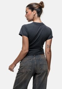 T-shirt noir ajusté à manches courtes, associé à un jean en denim délavé taille haute. Tatouages notables visibles sur le bras.