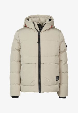 Person iført en beige dunjakke, mørke jeans og hvide sneakers, stående med hænderne i jakkelommerne.