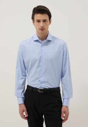 Jeune homme portant une chemise bleu clair boutonnée rentrée dans un pantalon noir avec une ceinture noire, debout devant un fond uni.