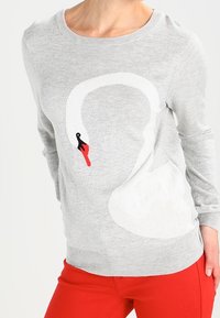 Pull gris avec un grand motif de cygne blanc arborant un bec rouge et noir, porté avec un pantalon rouge vif.