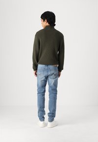 Olíva zöld bordázott pulóver magas gallérral, világoskék, slim fit farmerrel, hátsó zsebfelvarrással és fehér sportcipővel párosítva.