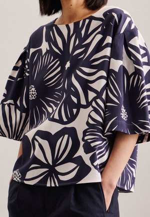 Femme portant un haut ample noir et blanc avec de grands motifs floraux et des manches courtes larges, les mains dans les poches d'un pantalon sombre.