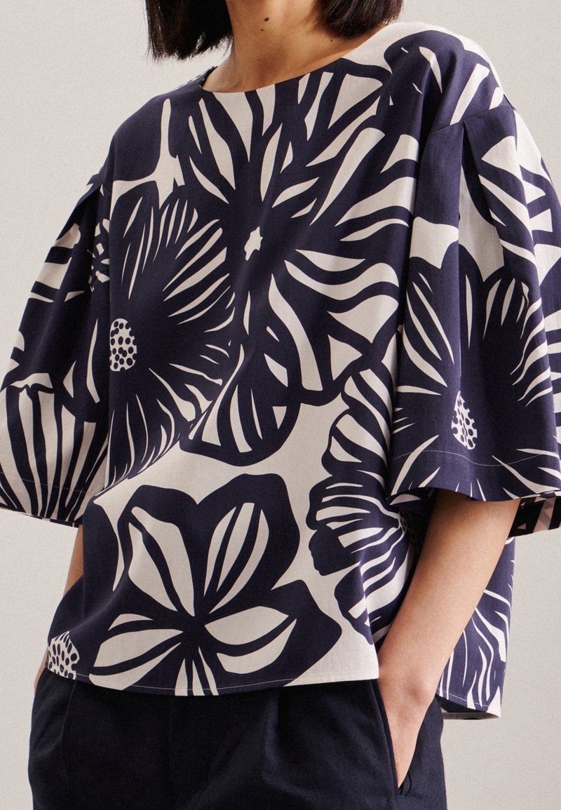 Femme portant un haut ample noir et blanc avec de grands motifs floraux et des manches courtes larges, les mains dans les poches d'un pantalon sombre.