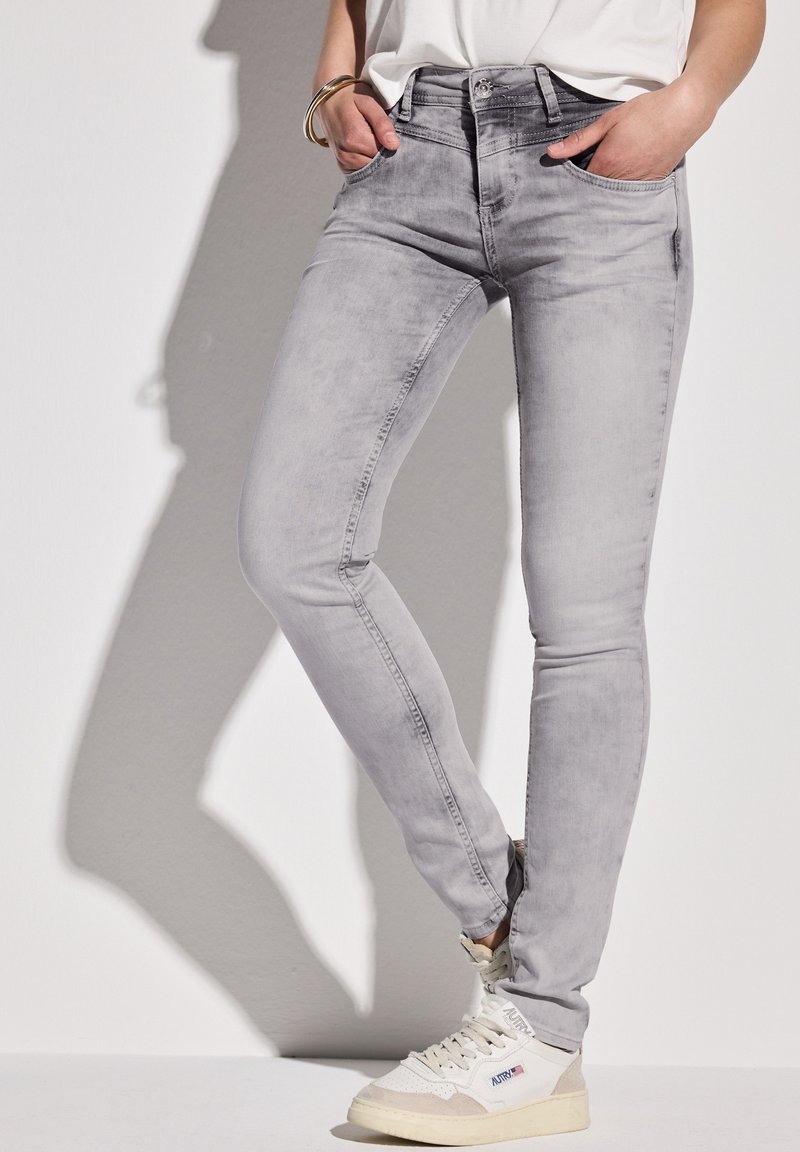 Personne portant un jean skinny gris clair, des baskets blanches avec logo, les mains dans les poches, debout contre un mur blanc projetant une ombre.