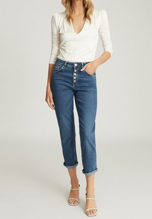 Vrouw met een witte, lange mouwen, V-hals top, blauwe jeans met hoge taille en knoopsluiting, opgerolde pijpen en beige sandalen met bandjes, staand tegen een effen achtergrond.