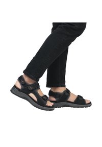 Sandales noires avec des sangles réglables, semelle en caoutchouc et détails perforés. Portées avec un jean noir, mettant en avant le confort et le soutien.