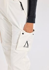 Vita cargobyxor med sidficka som har en tryckknapp, svart logotyp som säger "SLADE", och textilstruktur.