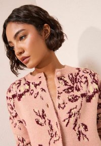 Gaiši rozā adīta kardigāna ar ziedu rakstiem tumši burgundzilā un dzeltenā krāsā, apaļu kakla izgriezumu un pogu aizdari. Mīksta tekstūra.