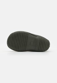Semelle de chaussure en caoutchouc verte avec un motif texturé et des rainures, featuring un emblème rectangulaire près du talon. Design plat et large.