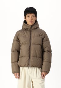 Brown puffer jacket with a hood, featuring a front zipper, stitched detailing, and a puffy texture. Worn over light-colored trousers.