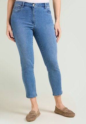 Frau trägt hellblaue, kurz geschnittene, enge Jeans und beigen Wildleder-Loafers, steht vor einem schlichten hellen Hintergrund.