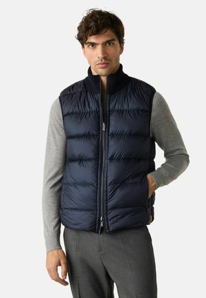 Mann trägt eine marineblaue gesteppte ärmellose Weste über einem grauen Langarmshirt und grauen Hosen, steht mit einer Hand in der Tasche.