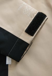 Beige and black jacket close-up; features a velcro strap, printed text, and smooth fabric texture with a color block design.
