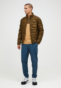 Giacca oliva verde morbida con una texture liscia, indossata sopra una camicia marrone chiaro, abbinata a pantaloni sportivi blu e scarpe nere e bianche.