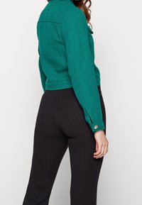 Teal strukturierte Jacke mit Kragen und einem Knopf am Manschette. Getragen mit schwarzen Nadelstreifenhosen. Sichtbare Nahtdetails auf der Rückseite.