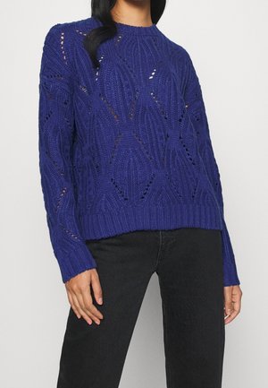 Vrouw draagt een paarse, getextureerde gebreide trui met een diamantpatroon en zwarte broek, staand met één hand ontspannen langs haar zij.