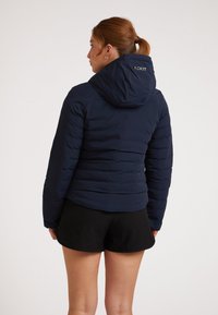 Giacca puffer navy con cappuccio, caratterizzata da una texture trapuntata e un design aderente. Include il marchio sulla parte superiore della schiena. Indossata con pantaloni corti neri.