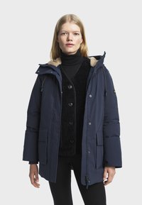 Marineblauwe pufferjas met een faux fur-gevoerde capuchon, grote voorzakken en een ritsluiting. Heeft een gestructureerde binnenvoering.