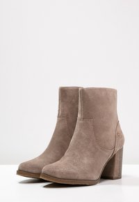 Botas de ante hasta el tobillo en un tono topo claro con punta redondeada, un tacón ancho alto y detalles de costura sutiles en los laterales.
