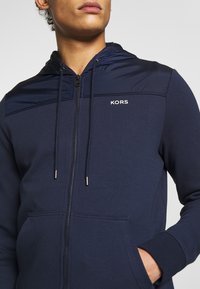 Sudadera con cierre de cremallera en color navy, con capucha, que presenta un panel de tela brillante en los hombros, bolsillos delanteros y un pequeño logo "KORS" en el pecho.