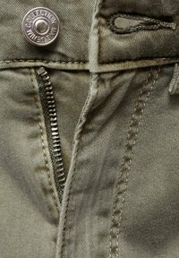 Maslinasto zelena denim tkanina s metalnim patentnim zatvaračem i dugmetom s ugraviranim natpisom "KOLEKCIJA". Vidljivo šivanje duž ivica.